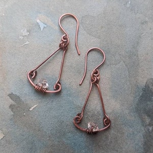Op de afbeelding: Een paar handgemaakte koperdraad oorbellen. Elke oorbel heeft een driehoekig frame met een kleine, heldere kristallen kraal gewikkeld in koperdraad aan de basis. De oorhaken zijn ook van koper, met decoratieve krullen aan de bovenkant.