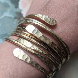 Op de afbeelding: Een set van zes gouden, gehamerde metalen armbanden. De armbanden zijn gestapeld op de pols van een persoon en hebben allemaal verschillende breedtes.