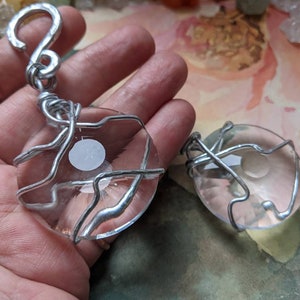 May include: Two clear, oval-shaped pendants with a white design in the center, wrapped in silver-colored wire. One pendant has a hook at the top. The pendants are displayed against a soft, colorful background.