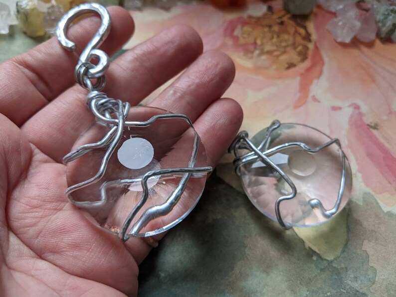 May include: Two clear, oval-shaped pendants with a white design in the center, wrapped in silver-colored wire. One pendant has a hook at the top. The pendants are displayed against a soft, colorful background.