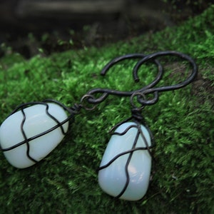 Può includere: Orecchini fatti a mano con pietre ovali azzurro pallido avvolte in filo metallico scuro. Gli orecchini pendono da un gancio curvo in metallo scuro. Lo sfondo è muschio verde vibrante.