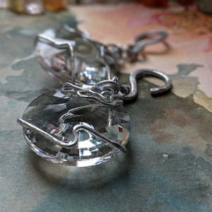 May include: Close-up of two clear crystal pendants with silver wire wrapping. The pendants have a faceted, circular shape, reflecting light. The silver wire is intricately wrapped around the crystals, creating a decorative design. The background is a soft, watercolor-style wash.