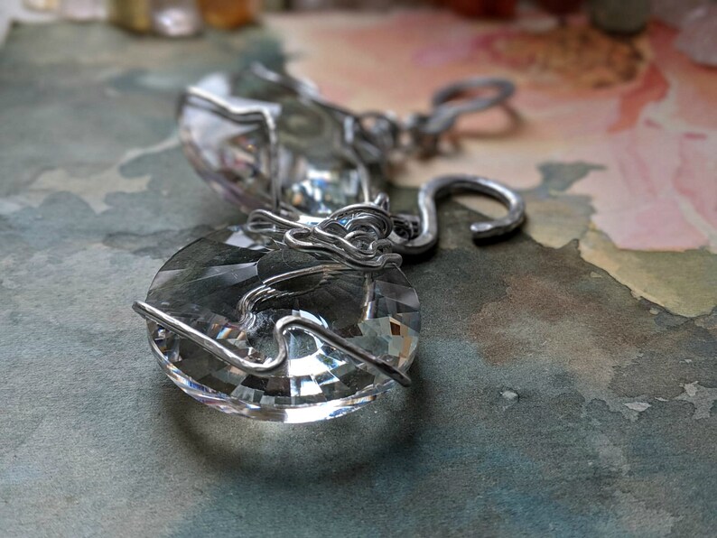 May include: Close-up of two clear crystal pendants with silver wire wrapping. The pendants have a faceted, circular shape, reflecting light. The silver wire is intricately wrapped around the crystals, creating a decorative design. The background is a soft, watercolor-style wash.