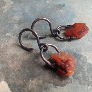 Könnte beinhalten: Handgefertigte Ohrringe mit grob geschnittenen, orange-roten Edelsteinen, die an dunklen Metallhaken hängen. Die Edelsteine sind unregelmäßig geformt und scheinen mit Draht befestigt zu sein. Die Ohrringe haben eine rustikale, natürliche Ästhetik.
