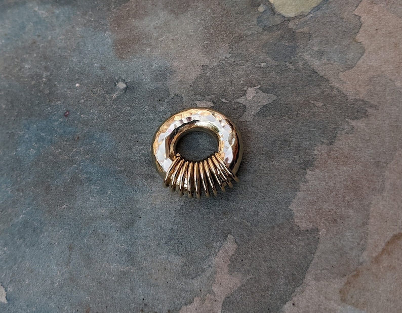 Puede incluir: Un colgante de anillo dorado con textura martillada y una serie de anillos m&aacute;s peque&ntilde;os apilados dentro del anillo m&aacute;s grande.