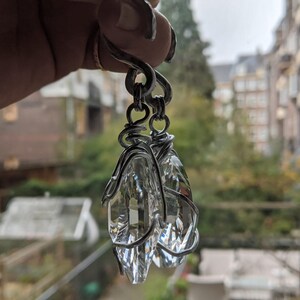 May include: A pair of clear crystal earrings with a silver wire wrap design. The earrings are suspended from a silver hook. The crystals are faceted, catching the light and creating a sparkling effect. The background is blurred, showing a window view.