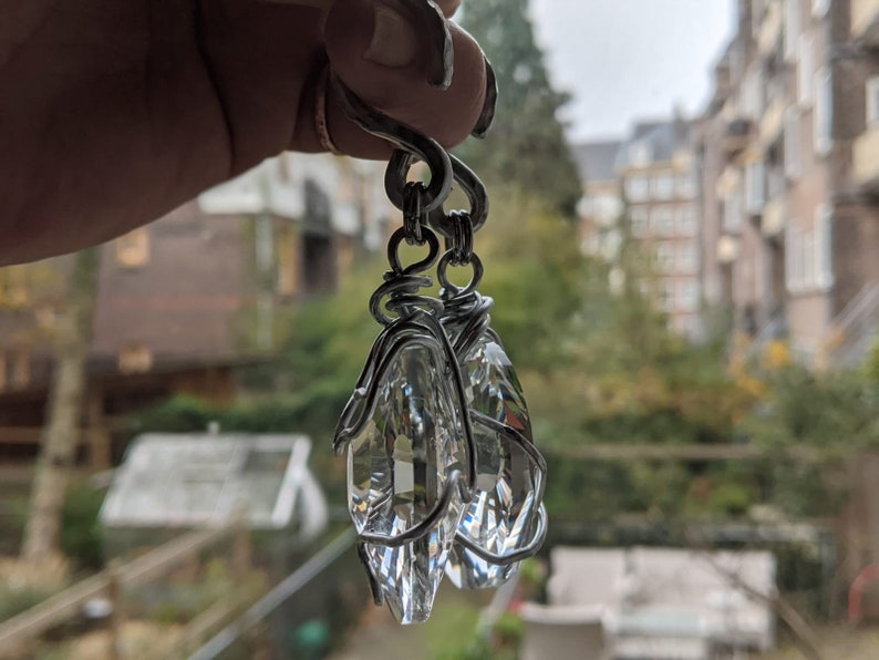 May include: A pair of clear crystal earrings with a silver wire wrap design. The earrings are suspended from a silver hook. The crystals are faceted, catching the light and creating a sparkling effect. The background is blurred, showing a window view.