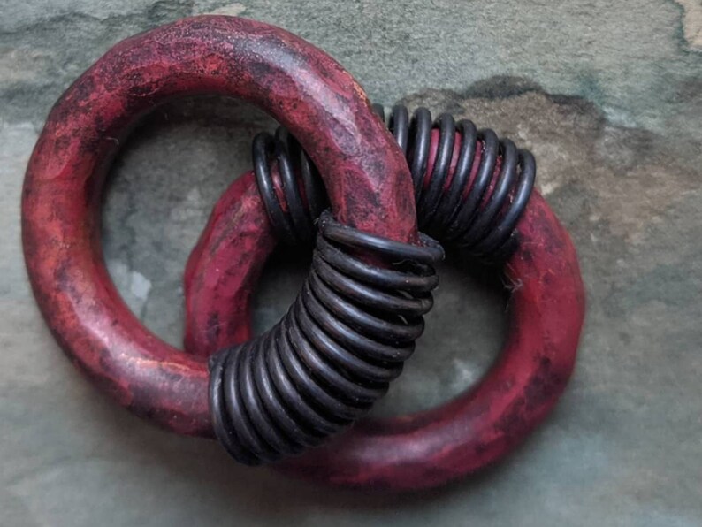 Puede incluir: Dos anillos de madera roja con una bobina de alambre negro envuelta alrededor de uno de los anillos. Los anillos est&aacute;n entrelazados.