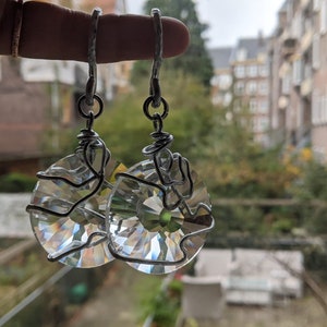 May include: A pair of clear crystal earrings with a circular design. The crystals are wrapped in silver-colored wire and hang from silver-colored hooks. The earrings are held up in front of a blurred background of trees and buildings.