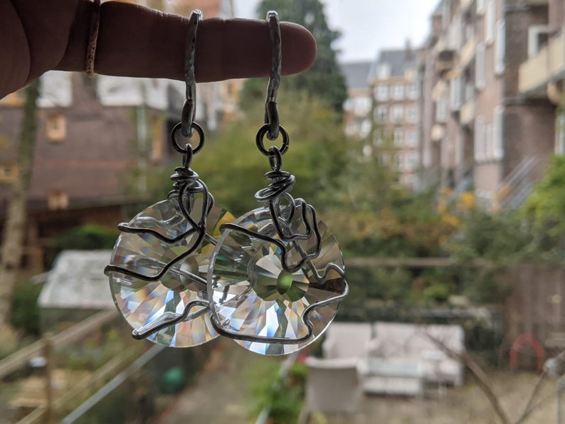 May include: A pair of clear crystal earrings with a circular design. The crystals are wrapped in silver-colored wire and hang from silver-colored hooks. The earrings are held up in front of a blurred background of trees and buildings.