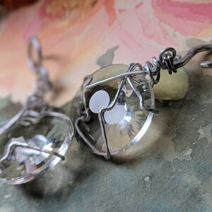 May include: Clear crystal earrings with a faceted, heart-shaped design, encased in silver-colored wire. The earrings have a hook closure. The background is a watercolor wash of pink, green, and blue.