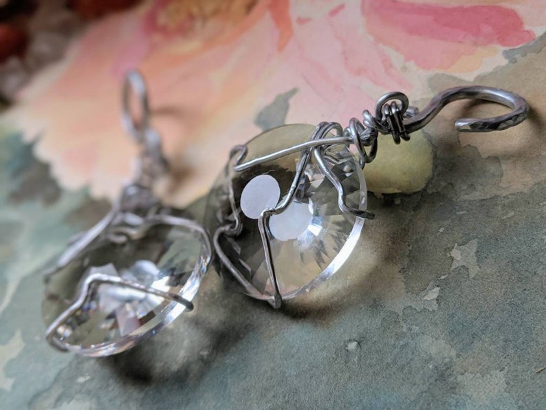 May include: Clear crystal earrings with a faceted, heart-shaped design, encased in silver-colored wire. The earrings have a hook closure. The background is a watercolor wash of pink, green, and blue.