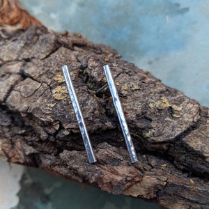 Op de afbeelding: Een paar zilveren oorstekers in staafvorm met een gehamerde textuur. De oorbellen worden gepresenteerd op een stuk getextureerd, bruin hout. Deze eenvoudige, elegante oorbellen zijn ongeveer 2,5 cm lang.