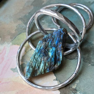 May include: A silver hammered metal bangle bracelet with a blue-green labradorite gemstone. The bracelet is made of multiple rings that are intertwined.