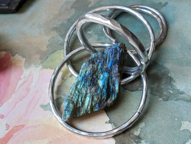 May include: A silver hammered metal bangle bracelet with a blue-green labradorite gemstone. The bracelet is made of multiple rings that are intertwined.