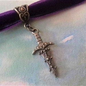 Könnte beinhalten: Ein lila Samt-Choker-Halskette mit einem silberfarbenen Dolch-Anhänger. Der Dolch hat aufwendige Details und hängt an einem dekorativen Verschluss. Der Hintergrund ist eine Aquarellwäsche aus Blau- und Grüntönen.
