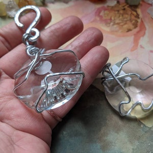 May include: Clear crystal pendants wrapped in silver wire. One pendant is attached to a silver hook. The crystals have a faceted design and a white design inside. The pendants are displayed on a hand.
