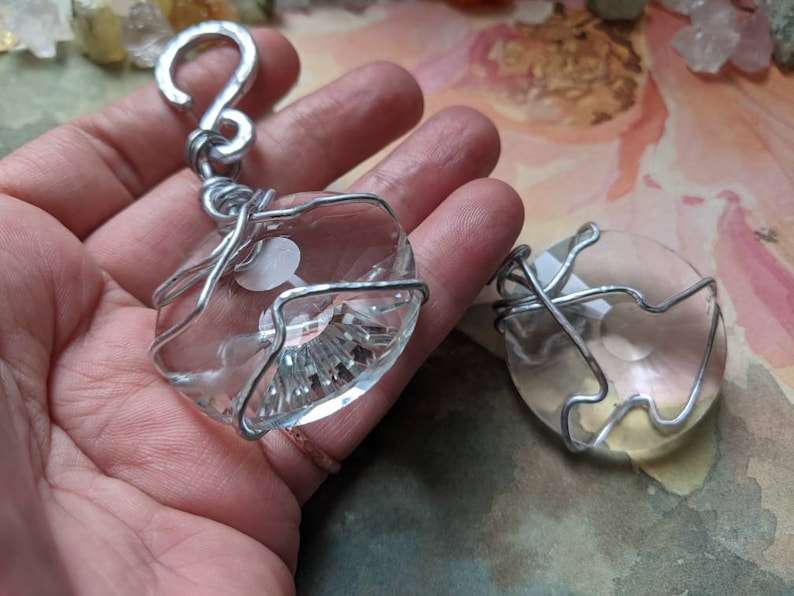 May include: Clear crystal pendants wrapped in silver wire. One pendant is attached to a silver hook. The crystals have a faceted design and a white design inside. The pendants are displayed on a hand.