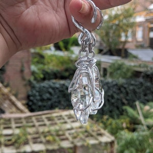 May include: A silver-colored, wire-wrapped pendant with two clear, faceted crystal teardrop-shaped stones. The pendant is suspended from a decorative loop. The background is a blurred view of greenery and buildings.