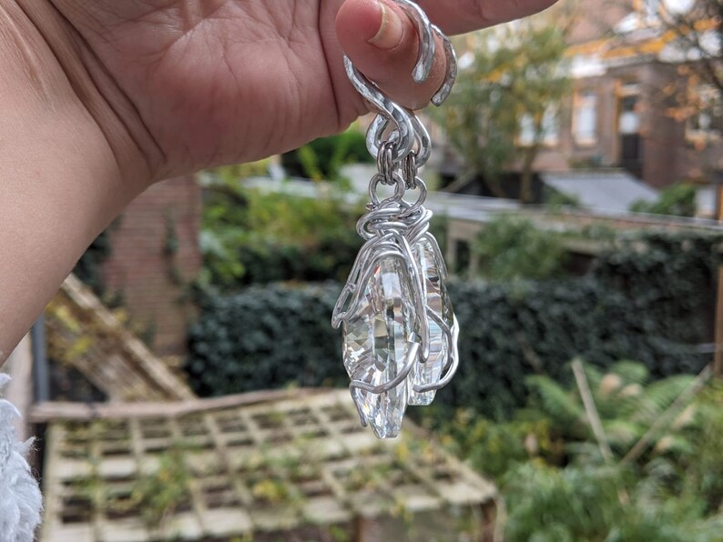 May include: A silver-colored, wire-wrapped pendant with two clear, faceted crystal teardrop-shaped stones. The pendant is suspended from a decorative loop. The background is a blurred view of greenery and buildings.