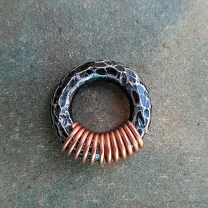 Op de afbeelding: Een zwarte en zilveren ring met een getextureerd oppervlak en een koperen draad eromheen gewikkeld.