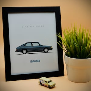 May include: A framed print featuring a teal Saab 900 Turbo car illustration. The print has the text "SAAB 900 TURBO" at the top and "SAAB" at the bottom. A small toy car and a potted plant are in the foreground.
