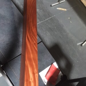 May include: A long, rectangular piece of wood with a rich reddish-brown color and a distinctive grain pattern. The wood is smooth and polished, and the grain is visible throughout the piece.