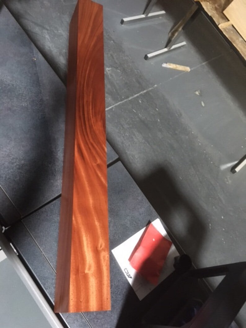 May include: A long, rectangular piece of wood with a rich reddish-brown color and a distinctive grain pattern. The wood is smooth and polished, and the grain is visible throughout the piece.