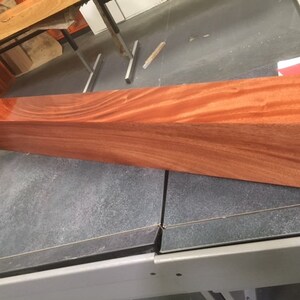 May include: A long, rectangular piece of polished wood with a reddish-brown color and a grain pattern. The wood is smooth and shiny.