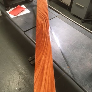 May include: A long, narrow piece of wood with a rich reddish-brown color and a beautiful grain pattern. The wood has been polished to a high sheen.