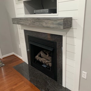 May include: A white fireplace mantel with a gray wood shelf and a black fireplace insert. The fireplace is surrounded by white shiplap walls.