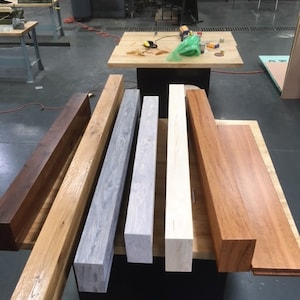 May include: Six wooden beams of different wood types and finishes. The beams are arranged in a row from left to right, with the darkest wood on the left and the lightest wood on the right. The beams are all about 12 inches long and 4 inches wide.