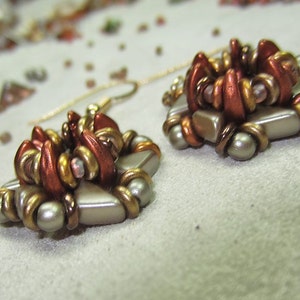 Könnte beinhalten: Ein Paar Ohrringe mit einem goldenen und kupfernen Blumendesign. Die Ohrringe bestehen aus kleinen Perlen und haben ein zartes, feminines Aussehen.