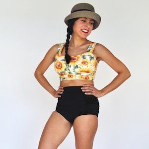 Op de afbeelding: Een vrouw draagt een zwart hoog getailleerd badpak met een zonnebloemprint top en een bruine hoed. Ze staat op een witte achtergrond en draagt bruine slippers.