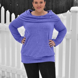 May include: A woman wearing a purple cowl neck sweater and black pants stands in front of a white picket fence. She is wearing black snow boots with fur trim.