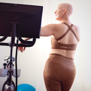 Puede incluir: Una persona con la cabeza rapada lleva un sujetador deportivo y leggings de leopardo marrón y negro, de pie frente a una bicicleta estática con una pantalla negra que dice "PELOTON".