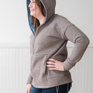 Puede incluir: Una sudadera gris con cremallera y capucha con forro a rayas azul y blanco. La sudadera tiene un bolsillo canguro y un corte holgado.