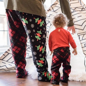 Può includere: Una persona e un bambino che indossano pantaloni da pigiama abbinati a quadri rossi e neri con un motivo a albero di Natale. La persona indossa una camicia verde e il bambino indossa una camicia rossa. Sono in piedi davanti a una tenda con un motivo bianco e nero.