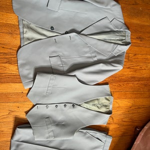 May include: A light grey three-piece suit comprising a blazer, waistcoat, and trousers. The blazer features two buttons and a notched lapel. The waistcoat has a five-button closure. The suit is made of a smooth fabric and is displayed on a wooden surface.