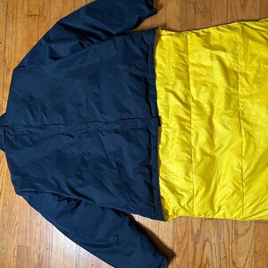 May include: A navy blue reversible jacket with a bright yellow interior. The jacket is laid out flat, showcasing the contrasting colors and quilted texture of the yellow side. The jacket appears to be made of a lightweight, possibly water-resistant material.