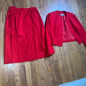 Könnte beinhalten: Ein leuchtend rotes Kostüm, bestehend aus einem knielangen Rock und einer passenden Jacke mit offenem Frontteil. Die Jacke hat lange Ärmel und eine taillierte Passform. Der Anzug ist aus einem einfarbigen, glatten Stoff gefertigt und wird auf einem Holzboden präsentiert.