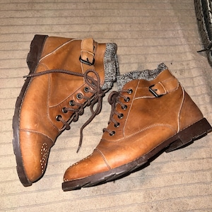 Op de afbeelding: Een paar bruine leren enkellaarsjes met donkerbruine zolen en veters. De laarzen hebben een gespsluiting en een grijs gebreid detail bij de enkel. De laarzen worden gepresenteerd op een getextureerd oppervlak.