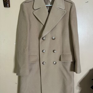 May include: A tan double-breasted overcoat with a notched collar and six buttons. The coat has two front pockets and epaulets on the shoulders. It is hanging on a white hanger against a neutral wall.