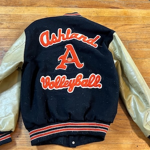 May include: Black and tan varsity jacket with red and white lettering. The back of the jacket features the words "Ashland Volleyball" and a large letter "A". The sleeves are tan leather.