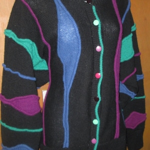May include: Black cardigan sweater with colorful accents. The sweater features a round neckline and button-up front. It has blue, purple, and teal decorative elements. The buttons are pink and teal. A unique, vintage style.