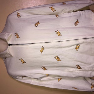 Puede incluir: Chaqueta blanca con cremallera y un patrón repetido de la palabra "AU" en amarillo.