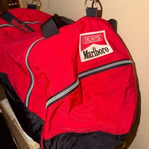 May include: A red and black duffel bag featuring a white rectangular patch with the text UNLIMITED Marlboro. The bag includes black nylon straps, silver-toned metal hardware, and a zippered closure. The material has a crinkled texture throughout.