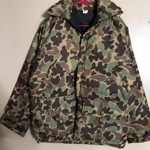 May include: Camouflage pattern jacket with a large collar and two front pockets. The jacket is in shades of brown, green, and tan. It is hanging on a red hanger.