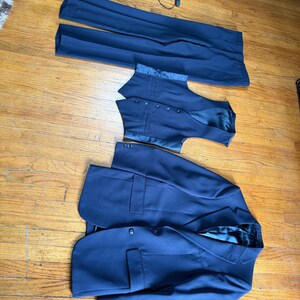 May include: A navy blue suit comprising trousers, a waistcoat, and a blazer. The suit features a fine pinstripe design. The blazer is buttoned and has pockets. The waistcoat has buttons. The suit is displayed on a wooden floor.
