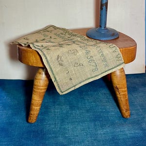 May include: A small, wooden stool with three legs supports a burlap cloth with green text and designs. A blue metal candle holder with a lit candle stands on the stool. The scene is set against a blue background.